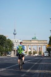 A man cycles down a bike lane on a summer day on Straße des 17 Juni in Berlin, Germany. The Brandenburg Gate and Berlin TV tower (Fernsehturm) are in the distance.