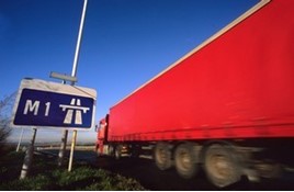 Red longer goods vehicle driving past M1 motorway sign