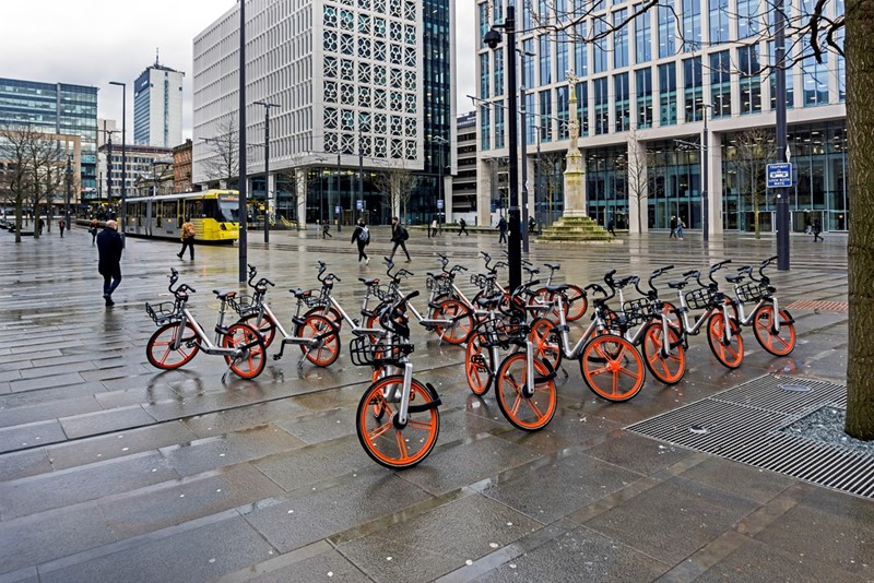  View of Manchester vity bikes. The bikes can be seen parked ready for use. A tram can be seen leaving the ttram station and people can be seen walking on the pavements.